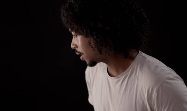 Kıvırcık saçlı genç Afro-Amerikalı adam siyah arka planda kameradan kafasını çeviriyor ve konuşuyor.