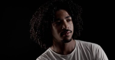 Siyah bir arka planda kameradan yüzünü çeviren, kıvırcık saçlı, düşünceli, Afro-Amerikan bir adamın portresi.