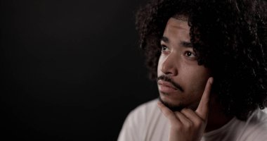 Düşünceli, kıvırcık saçlı Afro-Amerikalı bir adam çenesini tutuyor ve karanlık bir arka plana bakıyor.