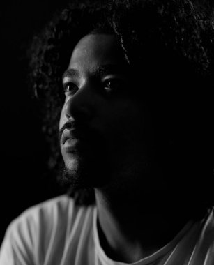 Kıvırcık saçlı, tek bir ışık kaynağıyla aydınlatılmış Afro-Amerikan genç bir adamın dramatik siyah-beyaz portresi.