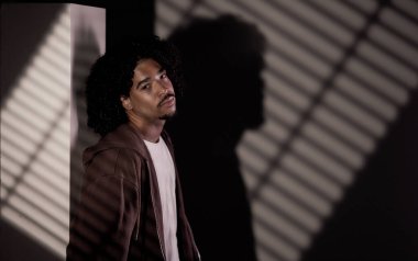 Şık, Afro-Amerikalı, kıvırcık saçlı, duvarın yanında çizgili gölgelerle poz veren genç bir adam.