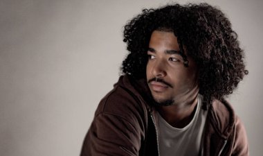 Tarafsız bir arkaplanda kameradan yüzünü çeviren kıvırcık saçlı, düşünceli, Afro-Amerikan bir adamın portresi.