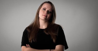 Uzun kahverengi saçlı, siyah tişörtlü, kollarını kavuşturup kendinden emin görünen güzel bir kadının stüdyo portresi.