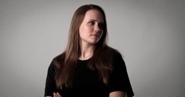 Uzun kahverengi saçlı, siyah tişörtlü, gri arka planda çapraz kolları olan ciddi bir genç kadının stüdyo portresi.