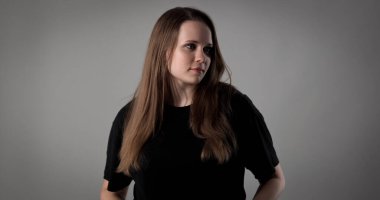 Uzun kahverengi saçlı, siyah tişörtlü, gri bir arka plana bakan genç bir kadının stüdyo portresi.