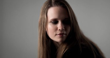 Gri bir arka plana bakan, uzun kahverengi saçlı, üzgün bir genç kadının portresi.