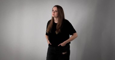 Elleri ceplerinde düşünceli genç bir kadın gri bir arka plana bakıyor.