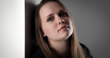 Uzun kahverengi saçlı, düşünceli ve huzurlu bir ifadeyle beyaz bir duvara yaslanmış beyaz bir kadının stüdyo portresi.