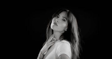 Uzun saçlı, beyaz tişörtlü ve boynuna dokunan güzel bir kadının siyah beyaz portresi.