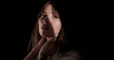 Hafif makyajlı güzel bir kadın, elleri boynunda, hafif bir ışıkla stüdyoda poz veriyor.