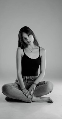 Genç bir kadın bacak bacak üstüne atmış kot pantolon ve siyah bluzla yerde oturuyor.