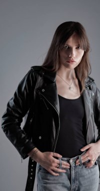 Uzun kahverengi saçlı, siyah deri ceketli, siyah bluzlu ve gri arka planda stüdyoda poz veren genç bir kadın.
