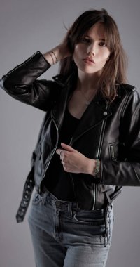 Uzun kahverengi saçlı, siyah deri ceketli ve kot pantolonlu genç bir kadın. Stüdyoda eli saçında poz veriyor.