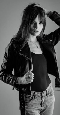 Uzun saçlı, deri ceketli ve kot pantolonlu şık bir genç bayan stüdyoda poz veriyor.