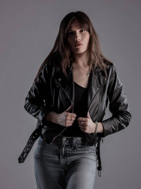 Uzun kahverengi saçlı, siyah deri ceketli ve gri arka planda poz veren genç bir kadın.