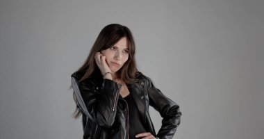 Uzun kahverengi saçlı, siyah elbiseli ve gri arka planda stüdyoda poz veren genç bir kadın.