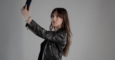 Genç bir kadın, gri arka planda siyah deri ceket giyip akıllı telefonuyla selfie çekiyor.