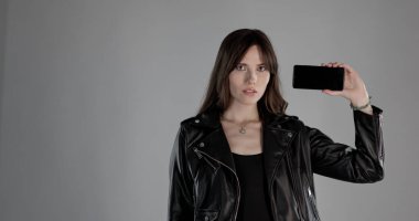 Beyaz ekranlı akıllı telefonu gösteren genç bir kadın gri arka planda siyah deri ceket giyiyor.