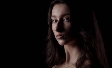 Uzun kahverengi saçlı, siyah arka planda poz veren güzel bir genç kadının stüdyo portresi.