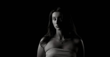 Uzun saçlı, çıplak omuzlu, kolyeli ve beyaz bluzlu, koyu arkaplandan çıkan genç bir kadının stüdyo portresi.