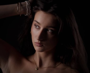 Altın kolye takan ve siyah arka planda bilezik takan eli saçları olan genç bir kadının stüdyo portresi.