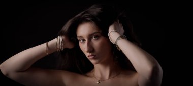 Güzel genç bir kadın uzun kahverengi saçlarını tutarken elleri bilezik takıyor ve siyah arka planda bir kolye takıyor.