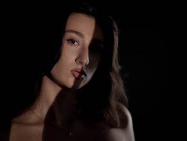 Uzun kahverengi saçlı ve çıplak omuzlu güzel bir genç kadının stüdyo portresi yüzünde gölgelerle dramatik bir ışıklandırma ile poz veriyor.