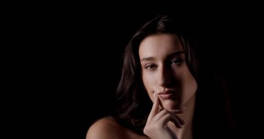 Stüdyoda uzun kahverengi saçlı, eli çenesinde ve koyu arkaplanda poz veren güzel bir genç kadın.