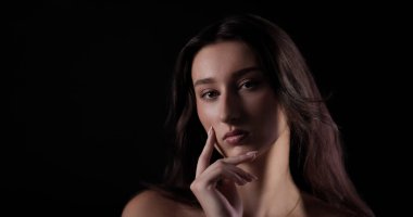 Uzun kahverengi saçlı genç bir kadının stüdyo portresi. Elleriyle çenesine dokunuyor. Koyu bir arka planda.