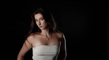 Uzun kahverengi saçlı, beyaz bluz ve kolye giyen, siyah arka planda poz veren güzel bir genç kadının stüdyo portresi.