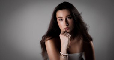 Uzun kahverengi saçlı, bilezik takan ve beyaz bluzlu eli çenesinde poz veren genç bir kadının stüdyo portresi.