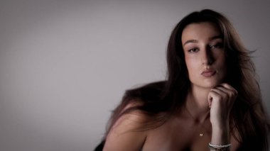 Uzun kahverengi saçlı, eli çenesinde poz veren, özgüven ve cazibe saçan güzel bir kadının stüdyo portresi.