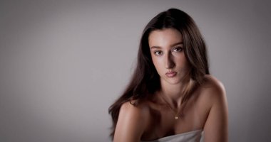 Uzun kahverengi saçlı, çıplak omuzlu, beyaz bluz ve altın kolye takan, gri arka planda poz veren güzel bir genç kadının stüdyo portresi.