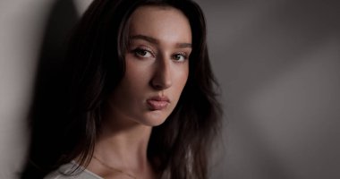 Uzun kahverengi saçlı ve ciddi ifadeli genç bir kadının stüdyo portresi.