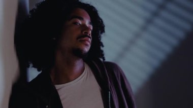 Dalgın, kıvırcık saçlı, loş ışıkta duvara yaslanmış genç bir Afro-Amerikalı.