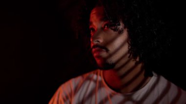 Yüzünde çizgili gölgeler olan kırmızı ve turuncu ışıkla aydınlatılmış kıvırcık saçlı genç bir Afro-Amerikalı 'nın dramatik portresi.