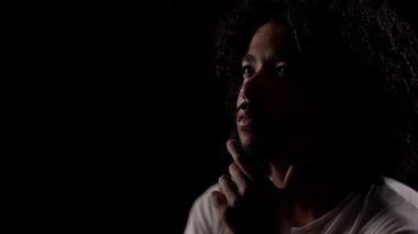 Karanlık bir odada çenesini tutarak dramatik ve düşünceli bir atmosfer yaratan kıvırcık saçlı düşünceli genç bir Afro-Amerikalı 'nın portresi.