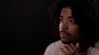 Düşünceli genç Afro-Amerikalı adam, elleri çenesinde, siyah arka planda izole edilmiş.