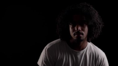 Kıvırcık saçlı ve sakallı Afro-Amerikalı genç bir adamın dramatik portresi, siyah arka planda hafif bir ışıkla aydınlatılmış.