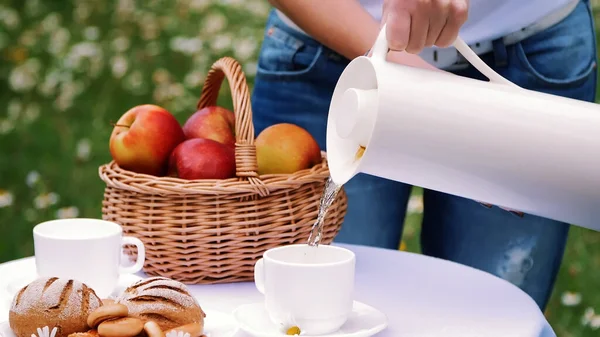在甘菊草坪的背景下 女人的手从一个白色的罐子里把茶倒进一个白色的杯子里 一个热水瓶 桌子上还有一个装有红苹果的篮子 高质量的照片 — 图库照片