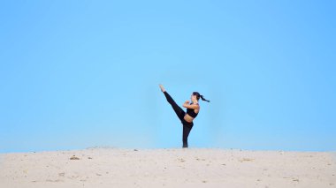 Siyah bluzlu atletik bir kız ve tayt, pratik tekmeleri, ayak tekmeleri, gölgelerle savaşmak, dövüşmek için antrenman. Issız bir sahilde, mavi gökyüzüne karşı, yazın, sıcak güneşin altında. Yüksek kalite