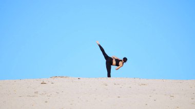 Siyah bluzlu atletik bir kız ve tayt, pratik tekmeleri, ayak tekmeleri, gölgelerle savaşmak, dövüşmek için antrenman. Issız bir sahilde, mavi gökyüzüne karşı, yazın, sıcak güneşin altında. Yüksek kalite