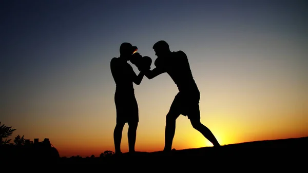 Two dark male figures, at sunrise, against the light, boxing, fighting in sparring, training in a pair of techniques of strikes. On sandy beach, in cargo port, near water, in summer. High quality