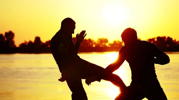 Two dark male figures, at sunrise, against the light, boxing, fighting in sparring, training in a pair of techniques of strikes. On sandy beach, in cargo port, near water, in summer. High quality
