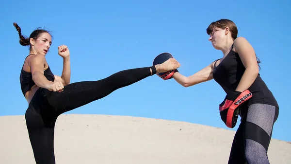 两个身穿黑色运动服的年轻女子 穿着一双运动服 在荒芜的海滩上 在炎热的阳光下 在蓝天的映衬下 练习踢腿 训练打斗 慢动作高质量的 — 图库照片