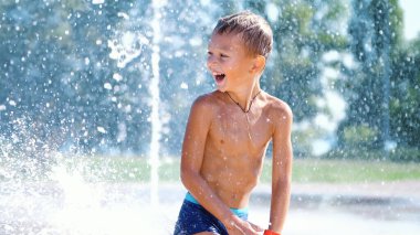 Yedi yıllık heyecanlı çocuk çeşmede, su jetleri arasında eğleniyor, etrafta koşuyor, serpiyor, eğleniyor, eğleniyor, sıcak bir yaz gününde. Şehirde yaz mevsimi. Ağır çekim. Yüksek kalite fotoğraf
