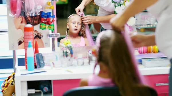 stock image Beautiful blond girl, of seven years old, braided two pigtails, do a hairstyle with pink locks of hair in a beauty salon, a hairdressers salon, in front of a large mirror. a little princess. High