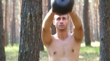 Çıplak, çıplak bir gövdesi olan genç atletik bir adam, çapraz formda olan ağırlık egzersizleri yapıyor. Ağır çekim. Çam ormanlarında, yazın, güneş ışınlarında.
