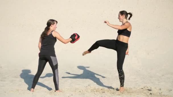 两个运动 年轻妇女在黑色健身服从事一双 工作出脚踢 在荒凉的海滩 蓝色的天空 在夏天 炎热的太阳下 — 图库视频影像