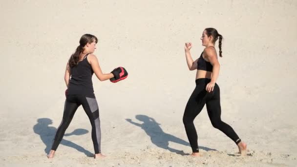 两个运动 年轻妇女在黑色健身服从事一双 工作出脚踢 在荒凉的海滩 蓝色的天空 在夏天 炎热的太阳下 — 图库视频影像